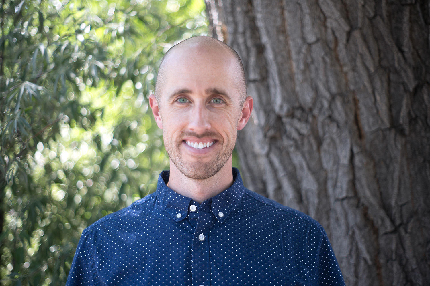 JCD director Garrett Stephens stands smiling in front of a tree in a dark blue collared shirt 