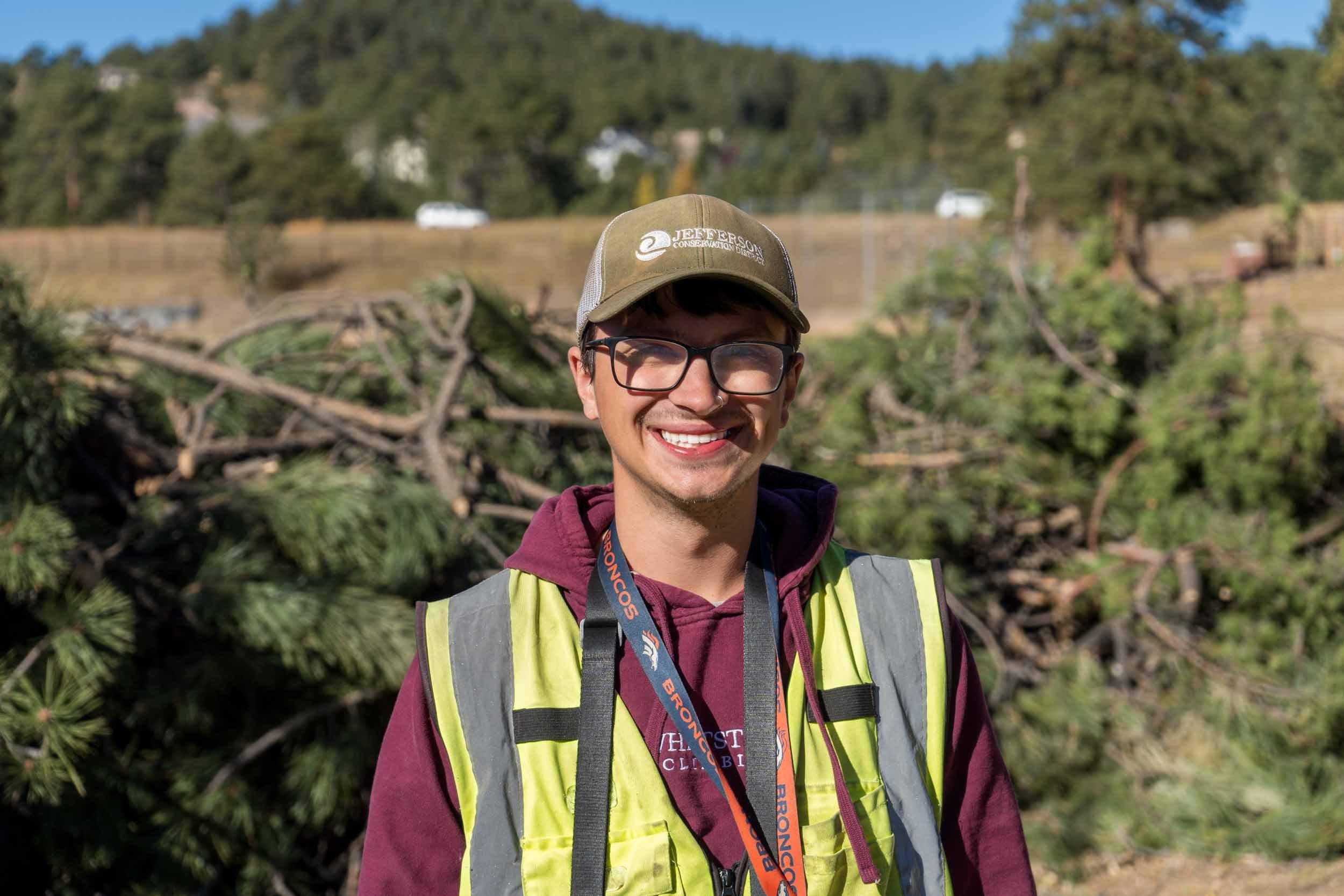 a man with glasses and a baseball hat stands in the sun with a yellow safety vest. He is smiling in front of piles of slash.