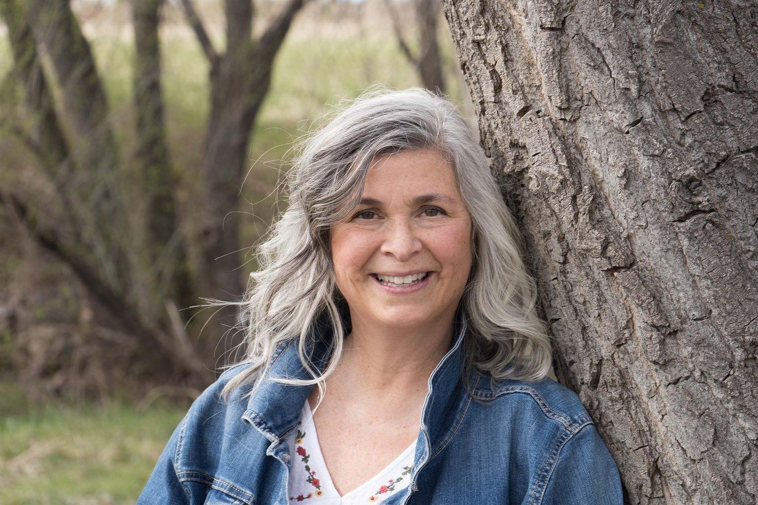 office manager Kim Blosser stands next to a tree smiling in a denim jacket