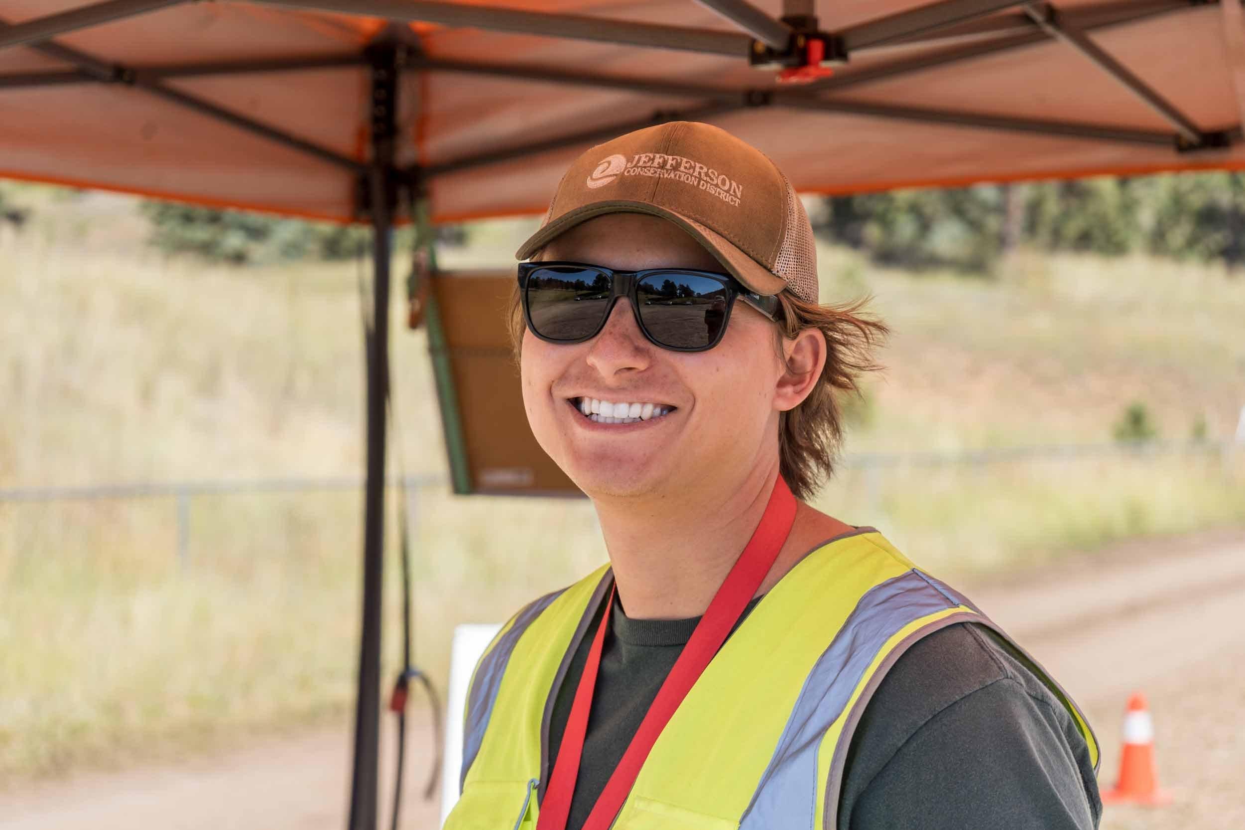 man smiling with sunglasses on, a baseball hat, and a safety vest