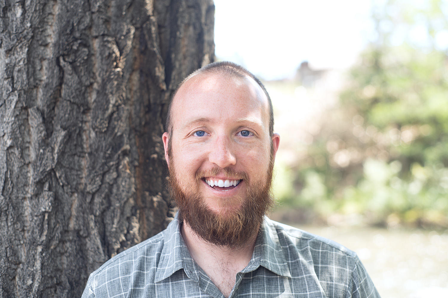 forest program manager Matt McLemore stands smiling in front of a tree in a gray plaid collared shirt